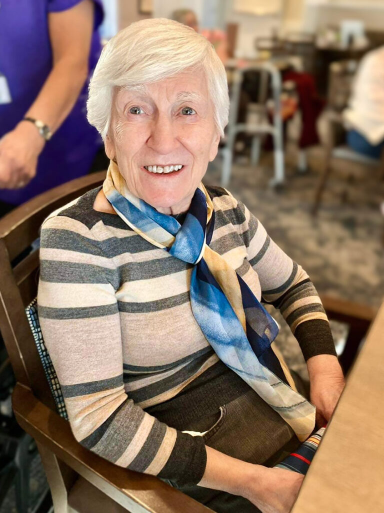 An older woman, Rose, sits in a chair, smiling warmly. She wears a gray and white striped long sleeve shirt with a blue and yellow scarf around her neck.