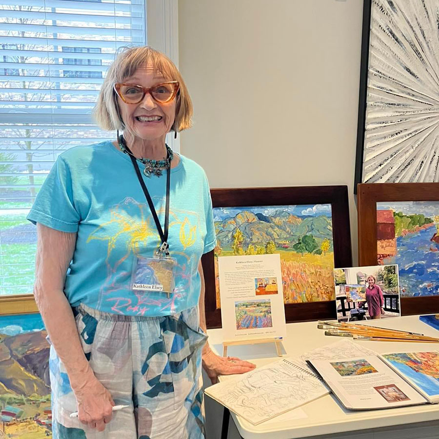 A talented elderly lady showcases her artwork, surrounded by a table adorned with various art materials.