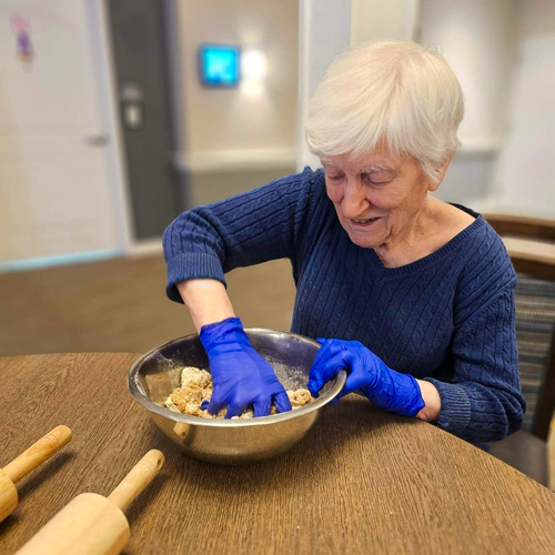 Senior woman with short white hair wearing blue gloves and a blue sweater, smiling and mixing dough in a metal bowl at a table with rolling pins in a senior living community.