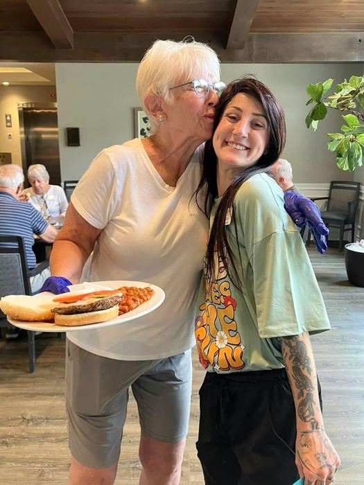 A senior lady holds a plate of food and gives a kiss on the cheek to a smiling staff member during a community event. Other residents are seen dining and enjoying the atmosphere in the background.