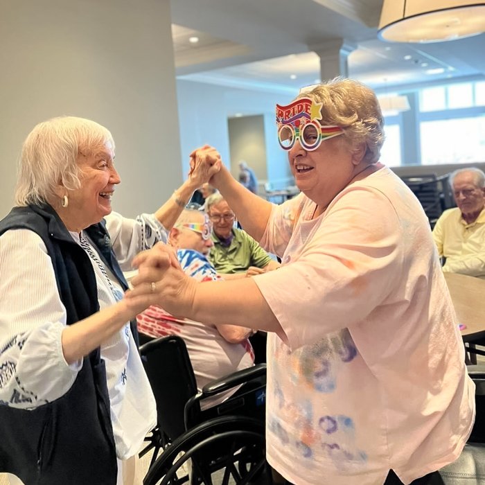 Two senior ladies dancing together during a community event. One of the ladies is wearing colorful 'PRIDE' glasses, and both are enjoying the moment with smiles on their faces, while other residents watch and participate in the background.