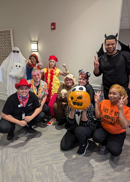Senior living employees dressed in creative Halloween costumes, including a ghost, cowboy, devil, and pumpkin mask, pose together celebrating a festive community event.