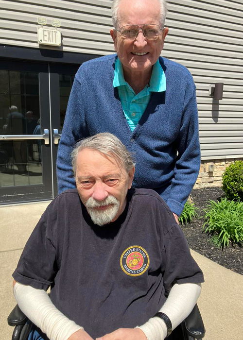 Two senior male residents smile together outside on a sunny day at a senior living community.