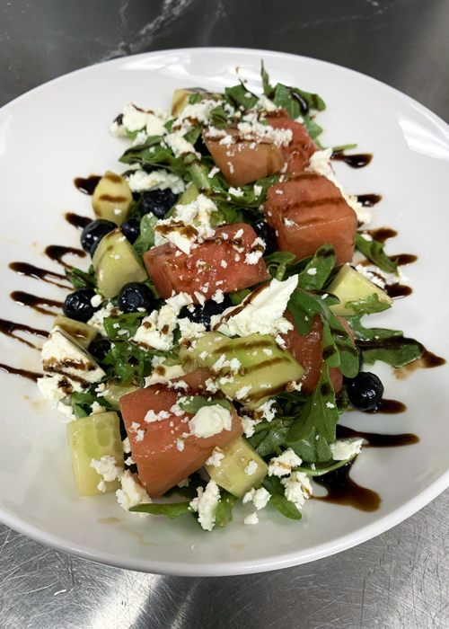 Fresh watermelon salad with feta, blueberries, cucumber, and arugula, drizzled with balsamic glaze and served on a white plate for a sweet and tangy flavor combo.