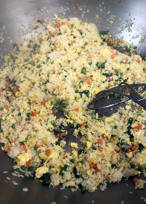 Colorful vegetable fried rice with scrambled eggs cooking in a large wok, featuring peas, carrots, corn, and spinach for a vibrant, quick stir-fry dish.