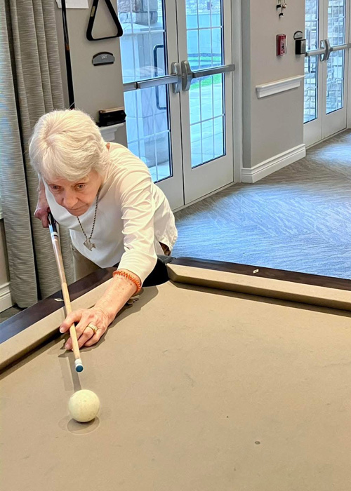Senior woman focused on her shot as she leans over a pool table, cue in hand, during a recreational event in a senior living space.