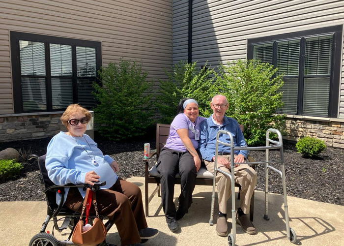 Two senior residents, one in a wheelchair and the other with a walker, sit outdoors with a smiling caregiver under the sunshine on a patio.