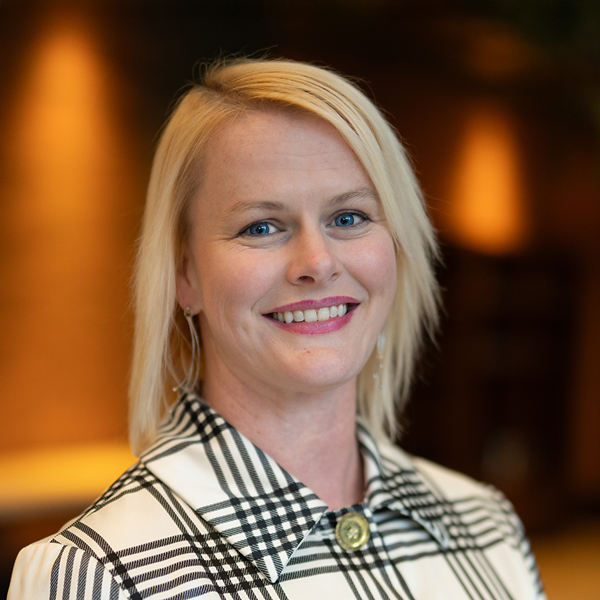 Headshot of Katie Metzger, Executive Director at Vitalia Stow, smiling in a black and white plaid-patterned collared dress shirt, with a softly lit background.