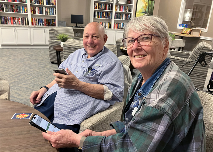 Two senior residents sit together in a cozy lounge, smiling while using their smartphones to explore the Arrow app.