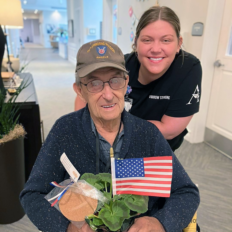A team member smiles next to a senior veteran holding a potted plant and an American flag.