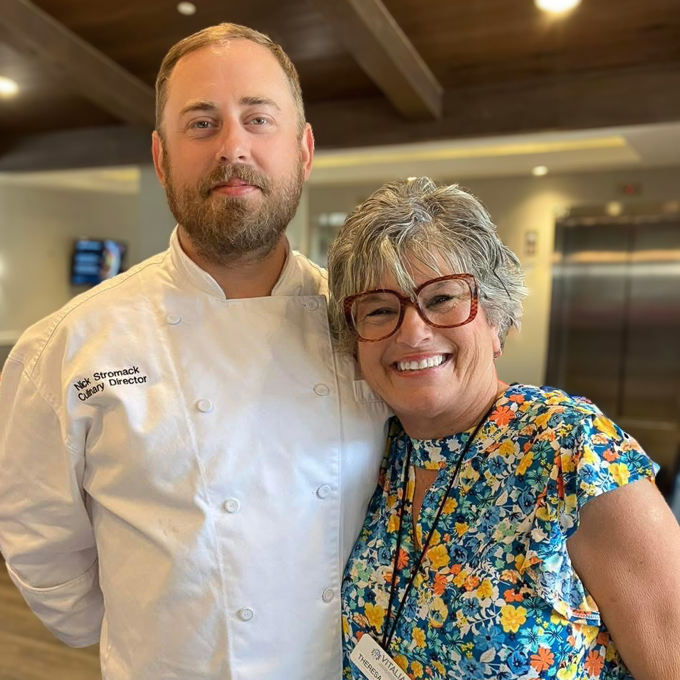Culinary Director Nick Stromack stands smiling with a woman in a floral top, capturing a warm and friendly moment inside a vibrant senior living community.