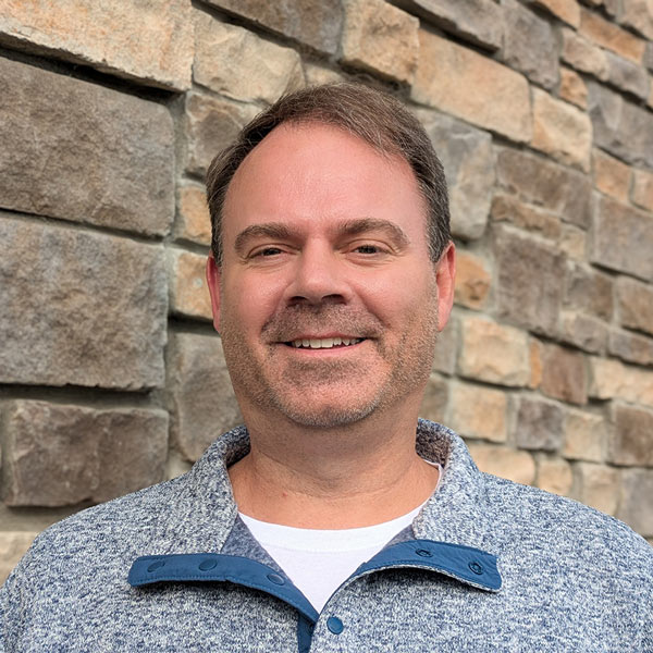 Keith Kasprzyk, Business Office Director at Vitalia Stow, smiling in a professional headshot. He has short brown hair, light stubble, and wears a blue and gray pullover with a white shirt underneath, standing in front of a stone wall background.