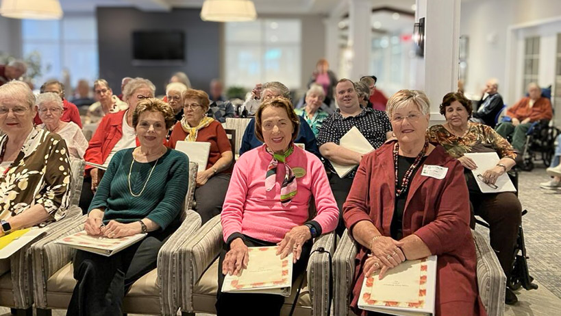 A large community gathering showing seated rows of smiling residents holding binders containing themed materials.