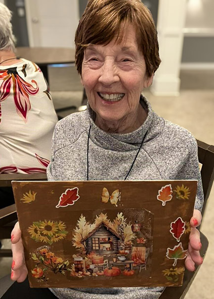 A senior woman smiles proudly, holding up a piece of artwork.