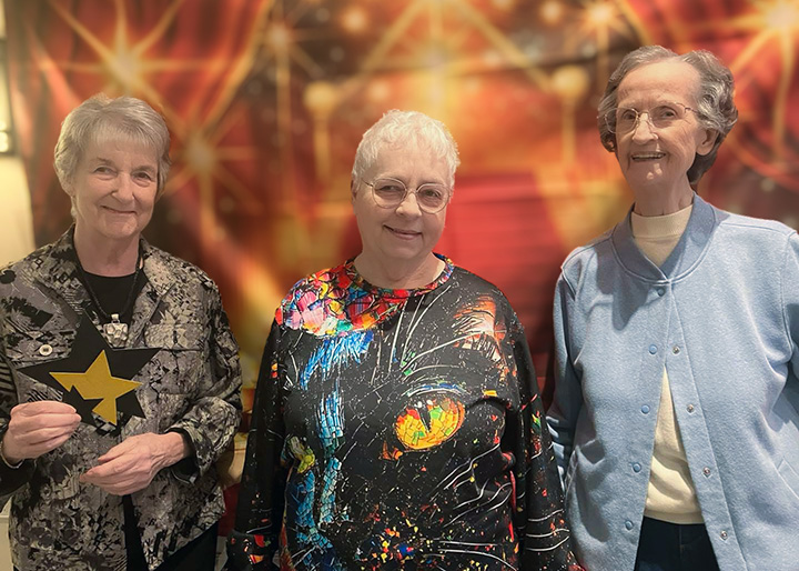 Three senior women smile in front of a fun backdrop at a community event.