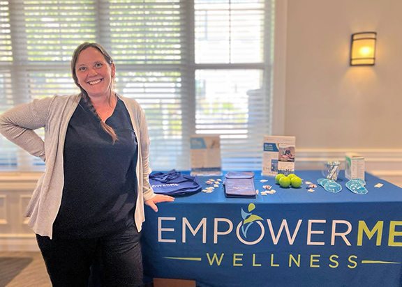 A woman smiles brightly beside a display table for EmpowerMe Wellness at an event.