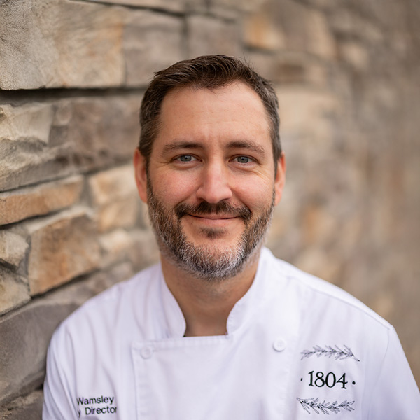 Christopher “Chris” Wamsley, Culinary Director at Vitalia Stow, smiling in a professional chef coat with short dark hair and a trimmed beard, standing against a stone wall background.