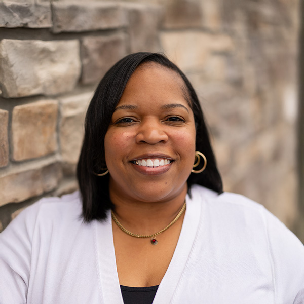 Dionna Henderson, Wellness Director at Vitalia Stow, smiling in a white cardigan with shoulder-length dark hair and gold hoop earrings, standing against a stone wall background.