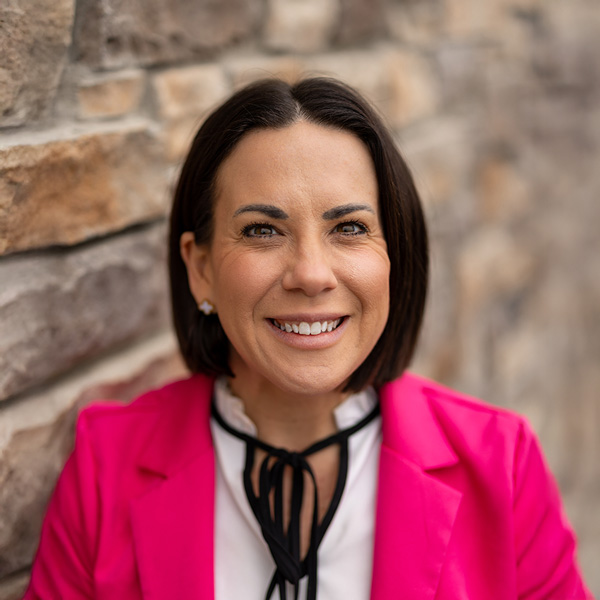 Heather Brenneman, Senior Living Director at Vitalia Stow, smiling in a professional headshot, wearing a bright pink blazer, with shoulder-length dark hair and a stone wall background.