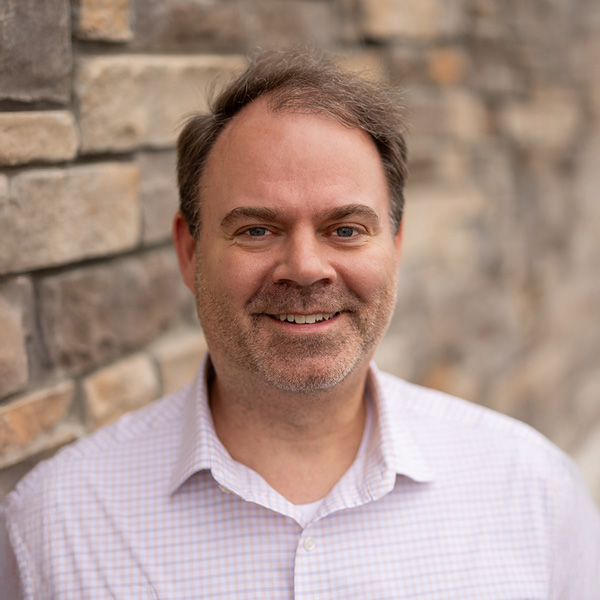 Keith Kasprzyk, Business Office Director at Vitalia Stow, smiling in a light checkered button-down shirt, standing against a stone wall with a softly blurred background.