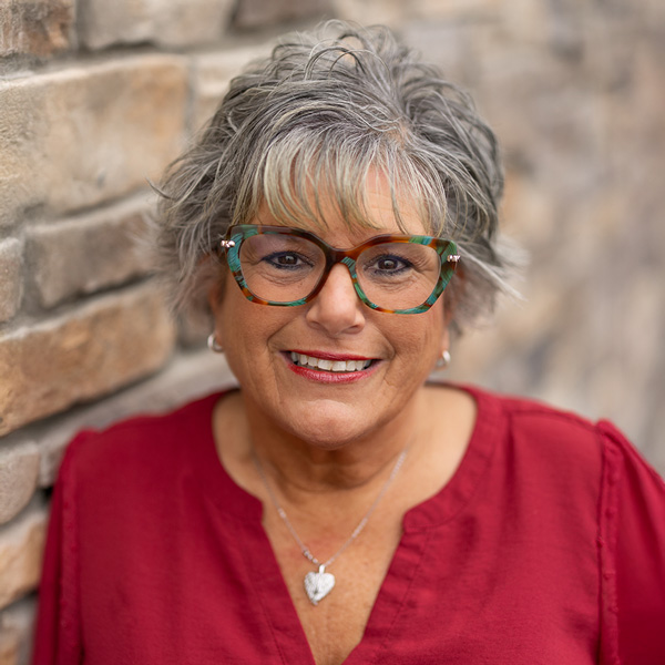 Theresa Erwin, Resident Services Director at Vitalia Stow, smiling warmly in a red blouse and patterned glasses, standing against a stone wall with a softly blurred background.