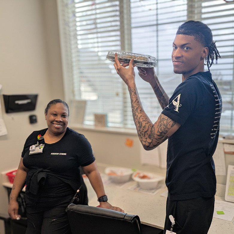 Two team members at Vitalia Stow smile, the man holding up a dessert.