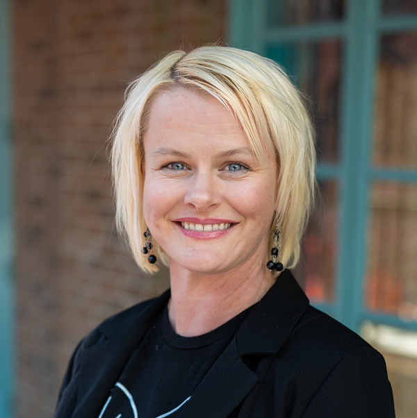 Katie Metzger, Executive Director at Vitalia Stow, smiling in a professional outdoor headshot with short blonde hair, blue eyes, black earrings, and softly blurred brick and teal window details in the background.
