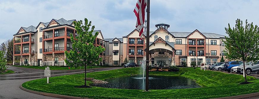 A wide shot of the exterior and grounds of Vitalia Stow, showcasing a small pond with fountain and the large, elegant exterior of the community.