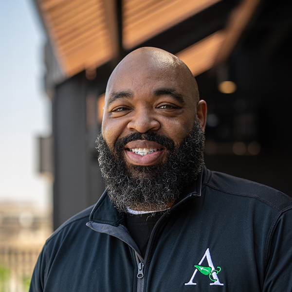 Thomas Drummer, Culinary Director at Vitalia Stow, smiles in an outdoor headshot taken under a shaded entrance, wearing a black jacket with the Arrow logo.