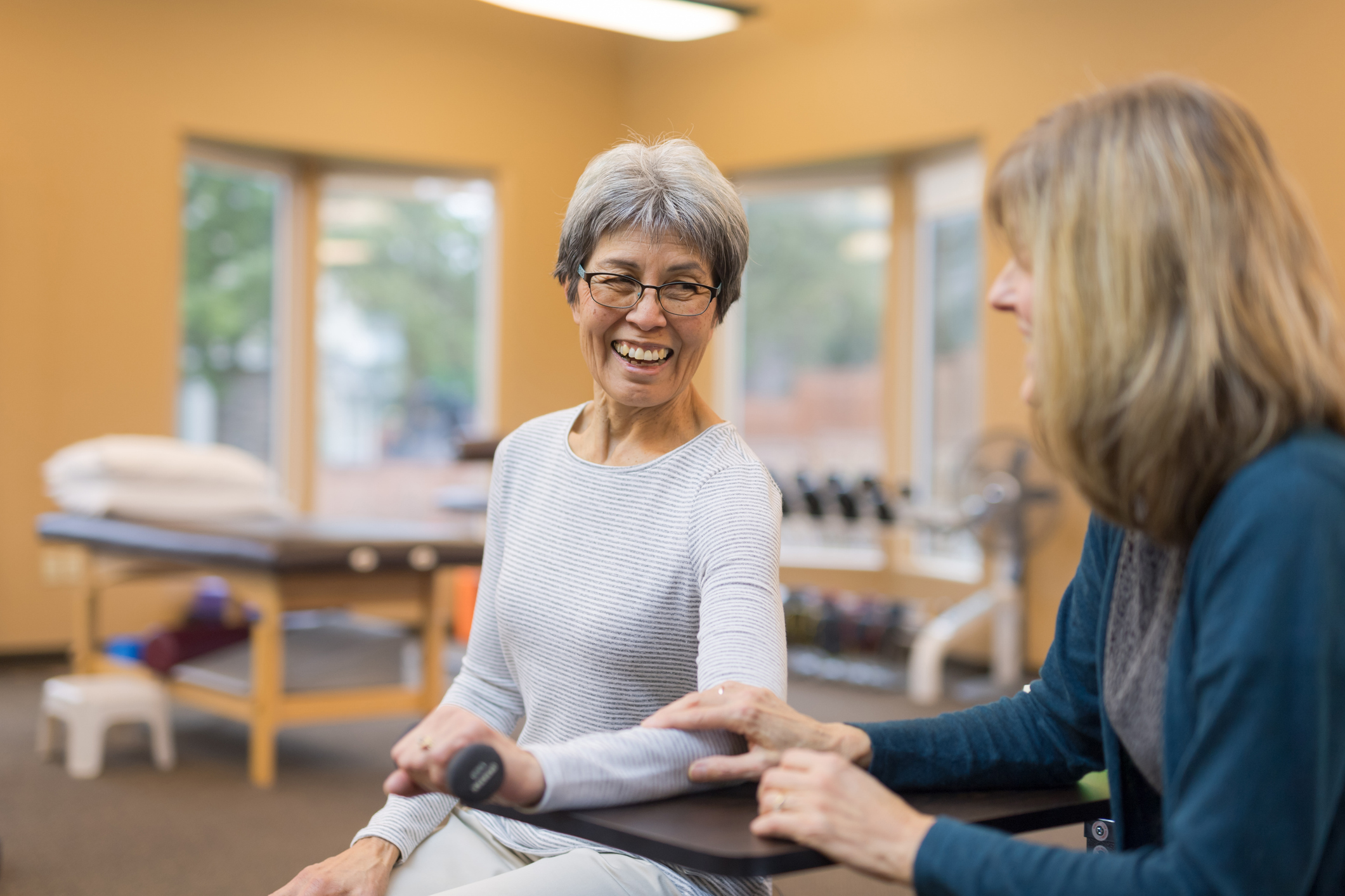 Older woman smiles while performing a seated arm exercise with a dumbbell as a fitness professional supports her movement.